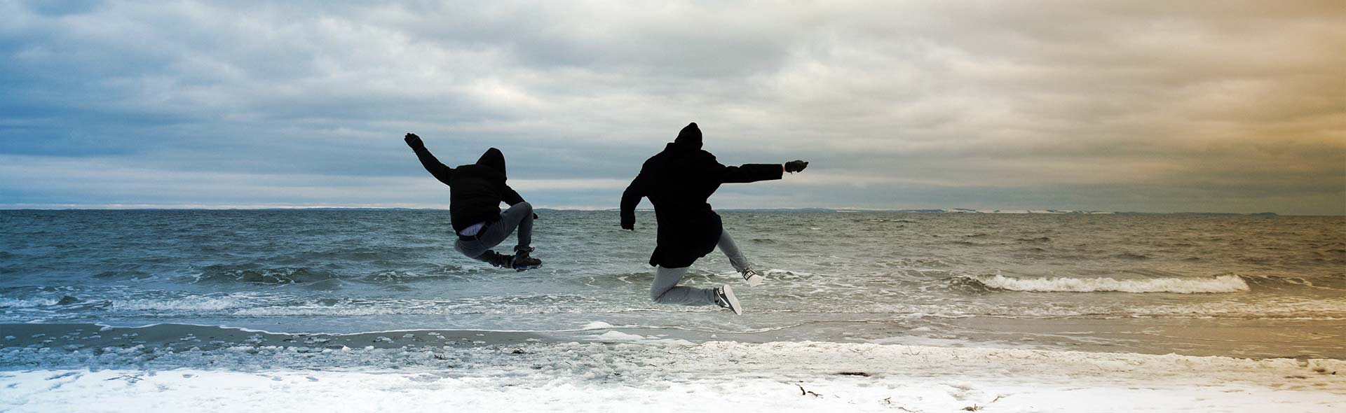 Redaktionsteam: Zwei junge Männer springen mit Wintermontur vor der Ostsee in die Höhee