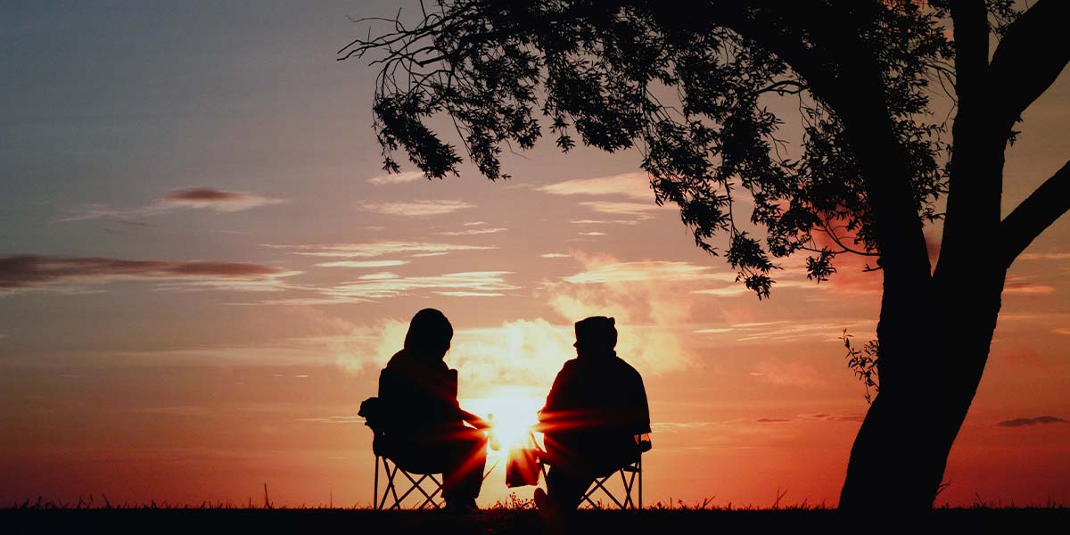 Zwei Personen sitzen vor einem Sonnenuntergang auf Campingstühlen und unterhalten sich